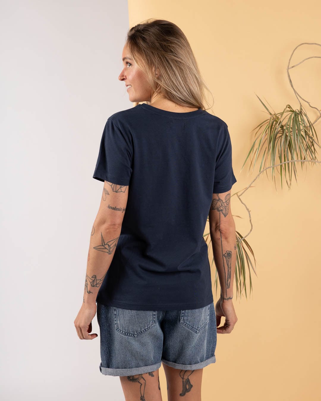 A tattooed person wears the Saltrock Celeste Stripe Women's Short Sleeve T-Shirt in blue, paired with denim shorts, standing with their back to the camera against a two-tone backdrop featuring a plant.