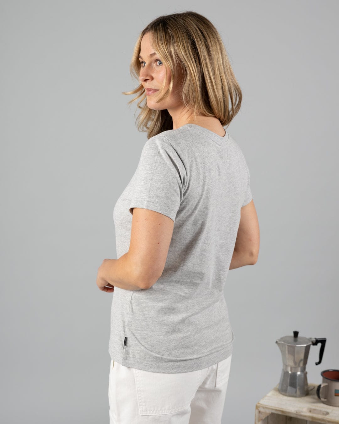 A blonde woman wears the Saltrock Velator Women's Short Sleeve T-Shirt in Grey Marl with white pants, standing slightly turned near a moka pot and cup on a small table.