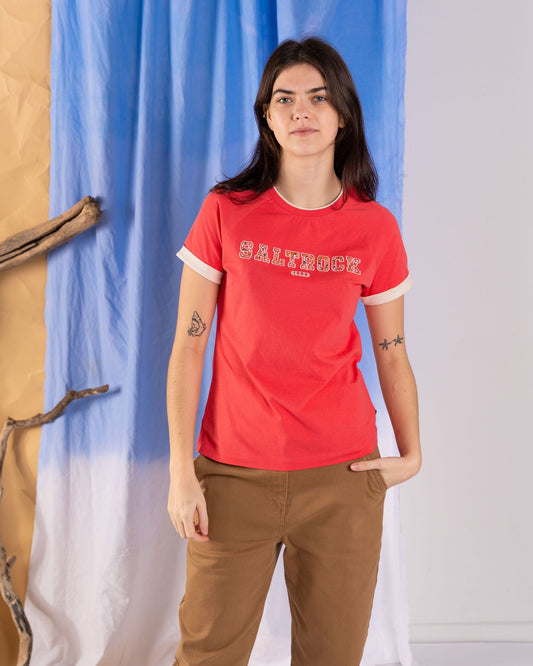 A woman with long dark hair wearing a Saltrock Campus Floral Women's Short Sleeve T-Shirt in coral and brown pants stands indoors near a blue and white fabric backdrop with wooden branches.