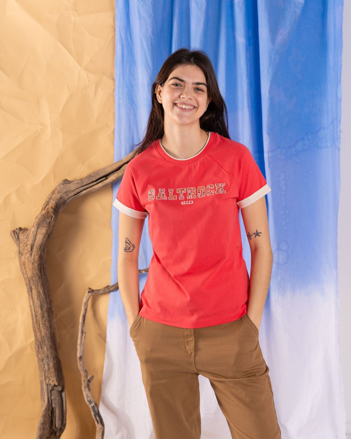 A person wearing a coral "Saltrock" Campus Floral women's short sleeve t-shirt and brown pants stands in front of a backdrop with blue, white, and beige sections, with pieces of driftwood beside them.