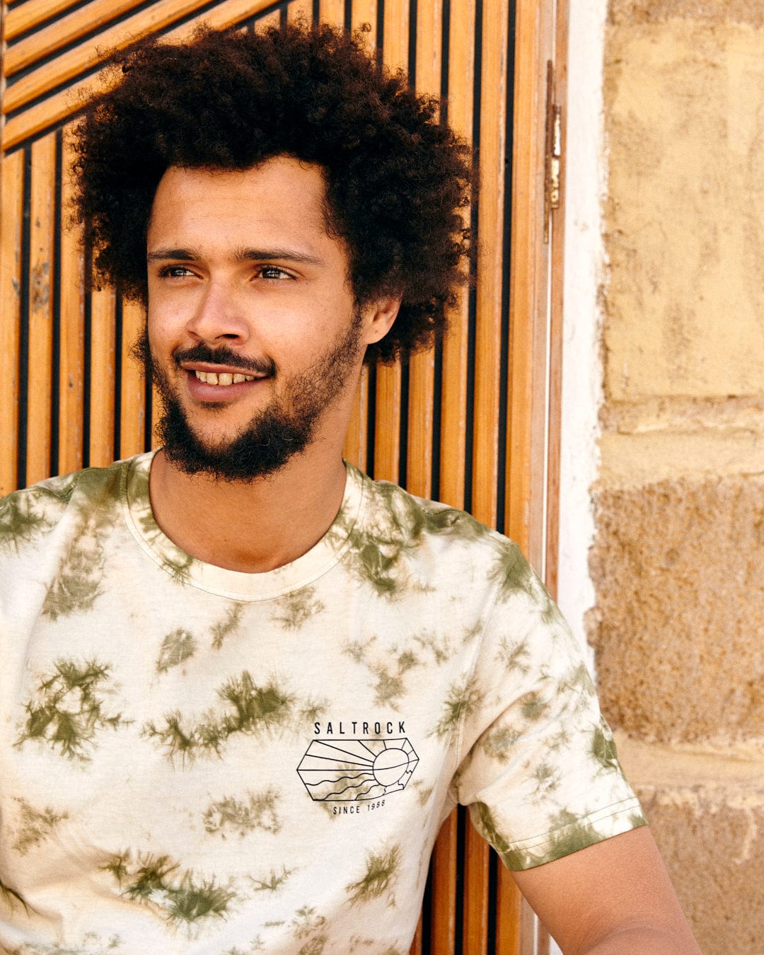 A young man with curly hair, wearing a tie-dye Saltrock Vantage Outline crew neck t-shirt in green, standing against a wall with wooden shutters.