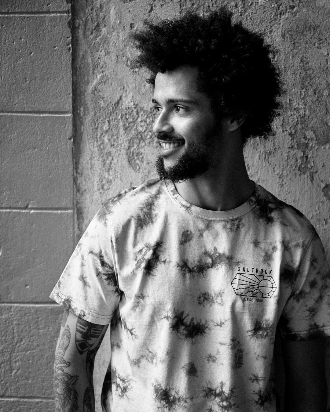A man with curly hair and a beard smiling in a Saltrock Vantage Outline - Mens Tie Dye T-Shirt - Green, standing against a textured wall, in a black and white photograph.