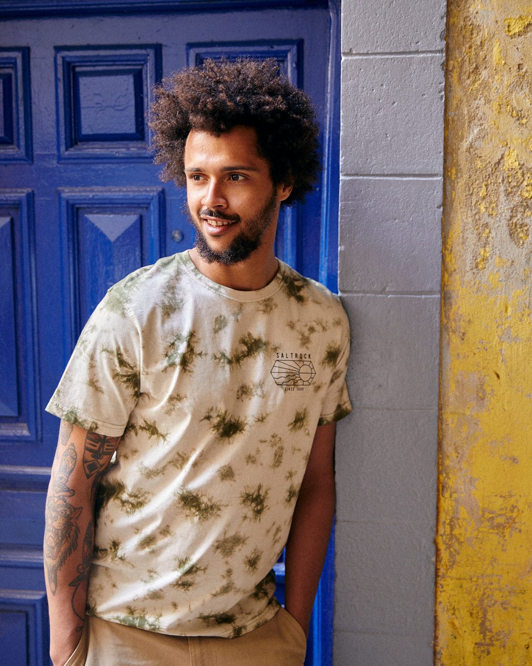 A young man with curly hair and a beard standing by a blue door, wearing a Saltrock Vantage Outline - Mens Tie Dye T-Shirt in Green and khaki pants, smiling slightly.