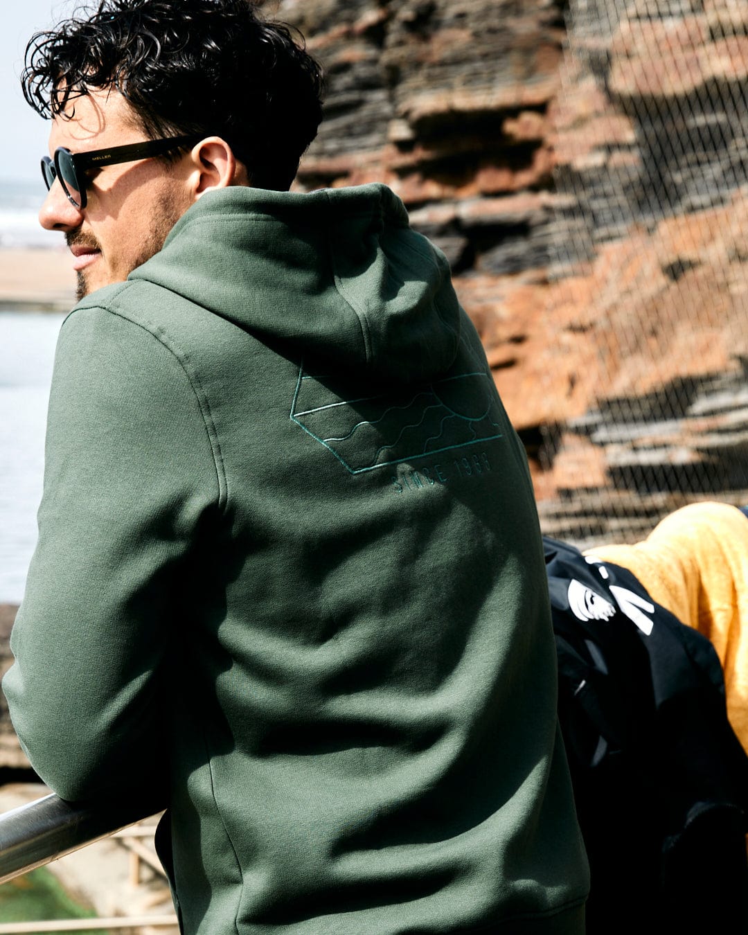 Man in Vantage Outline - Recycled Mens Zip Hoodie in Dark Green by Saltrock and sunglasses, looking away from the camera, standing by a railing with water and rocks in the background.
