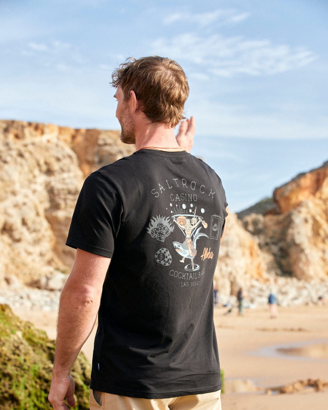 A man standing on a beach wearing a Saltrock Vegas Cocktail - Mens Short Sleeve T-Shirt in Black.