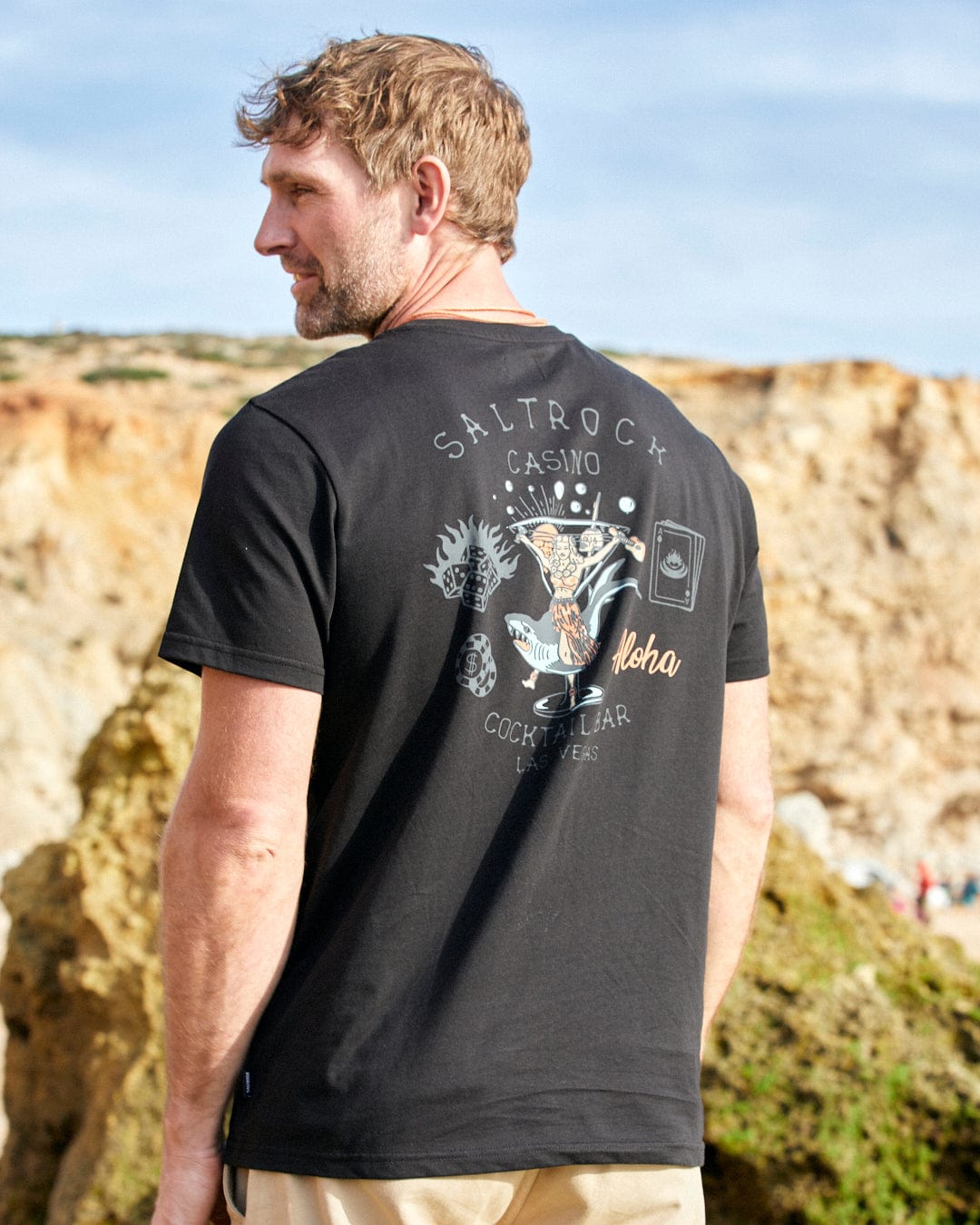 A man wearing a Saltrock Vegas Cocktail - Mens Short Sleeve T-Shirt in Black is standing on a rocky beach.