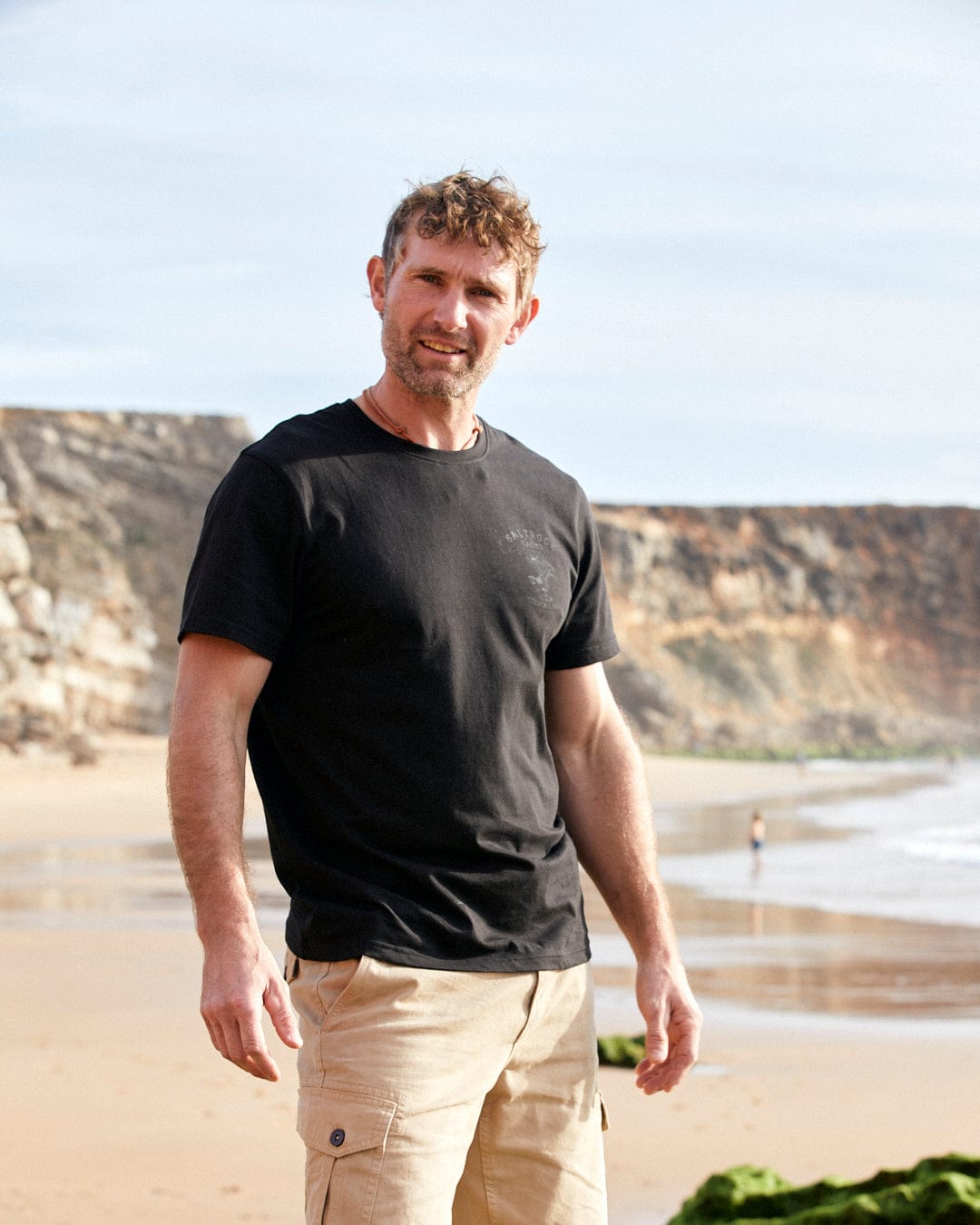 A man standing on a beach wearing a Vegas Cocktail - Mens Short Sleeve T-Shirt - Black made of Cotton by Saltrock.