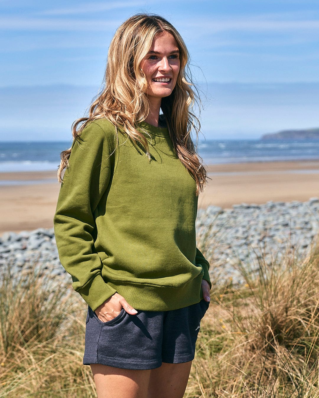 A woman wearing a Saltrock Velator - Womens Long Sleeve Sweat - Dark Green and shorts standing on the beach.