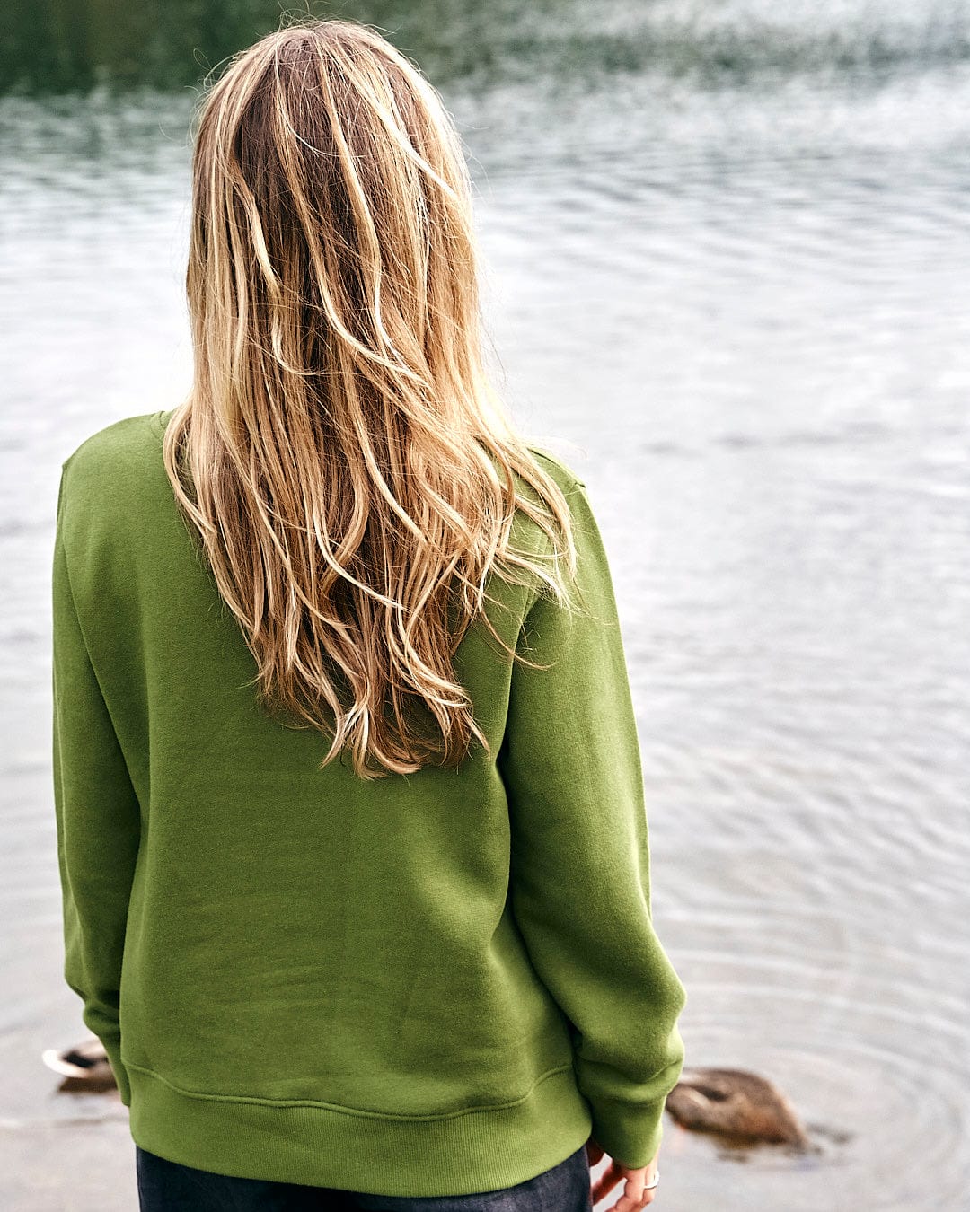 A woman wearing a Saltrock Velator Womens Long Sleeve Sweat in Dark Green standing by a lake.