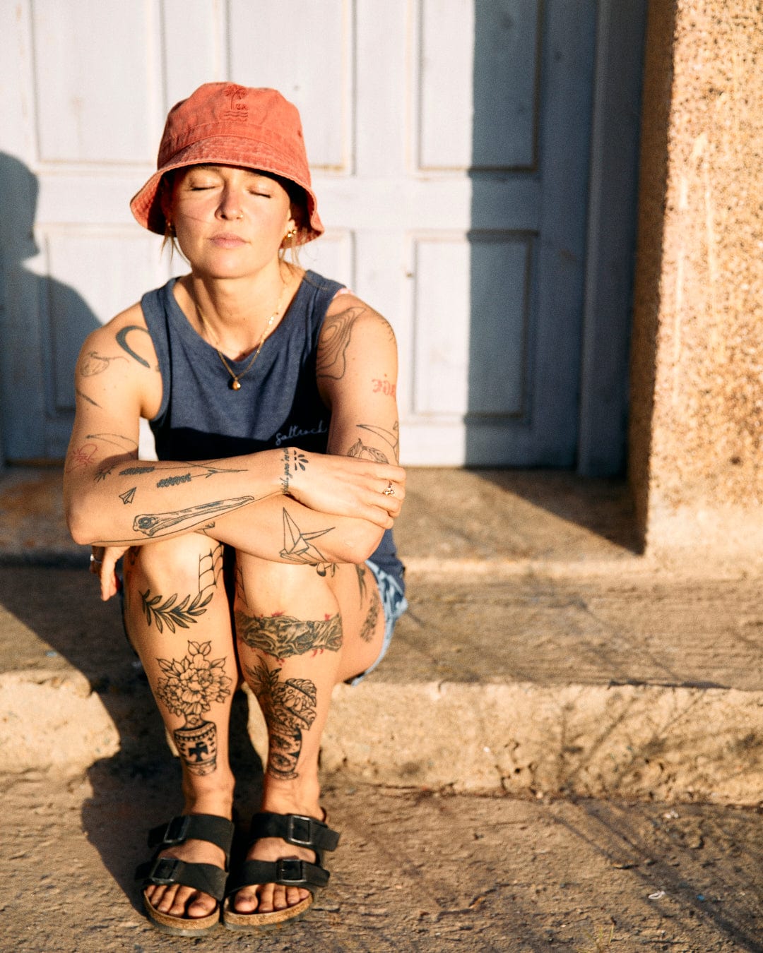 Woman with tattoos sitting in sunlight, eyes closed, wearing a red bucket hat and sandals with a crew neckline top, against a backdrop of a white door wearing the Saltrock Velator Womens Vest Blue.