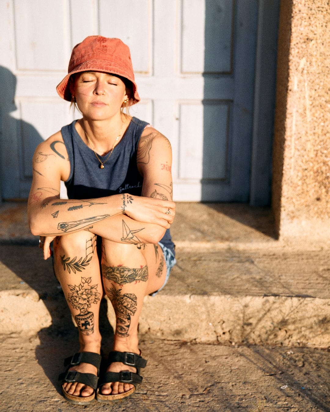 Woman with tattoos sitting in sunlight, eyes closed, wearing a red bucket hat and sandals with a crew neckline top, against a backdrop of a white door wearing the Saltrock Velator Womens Vest Blue.