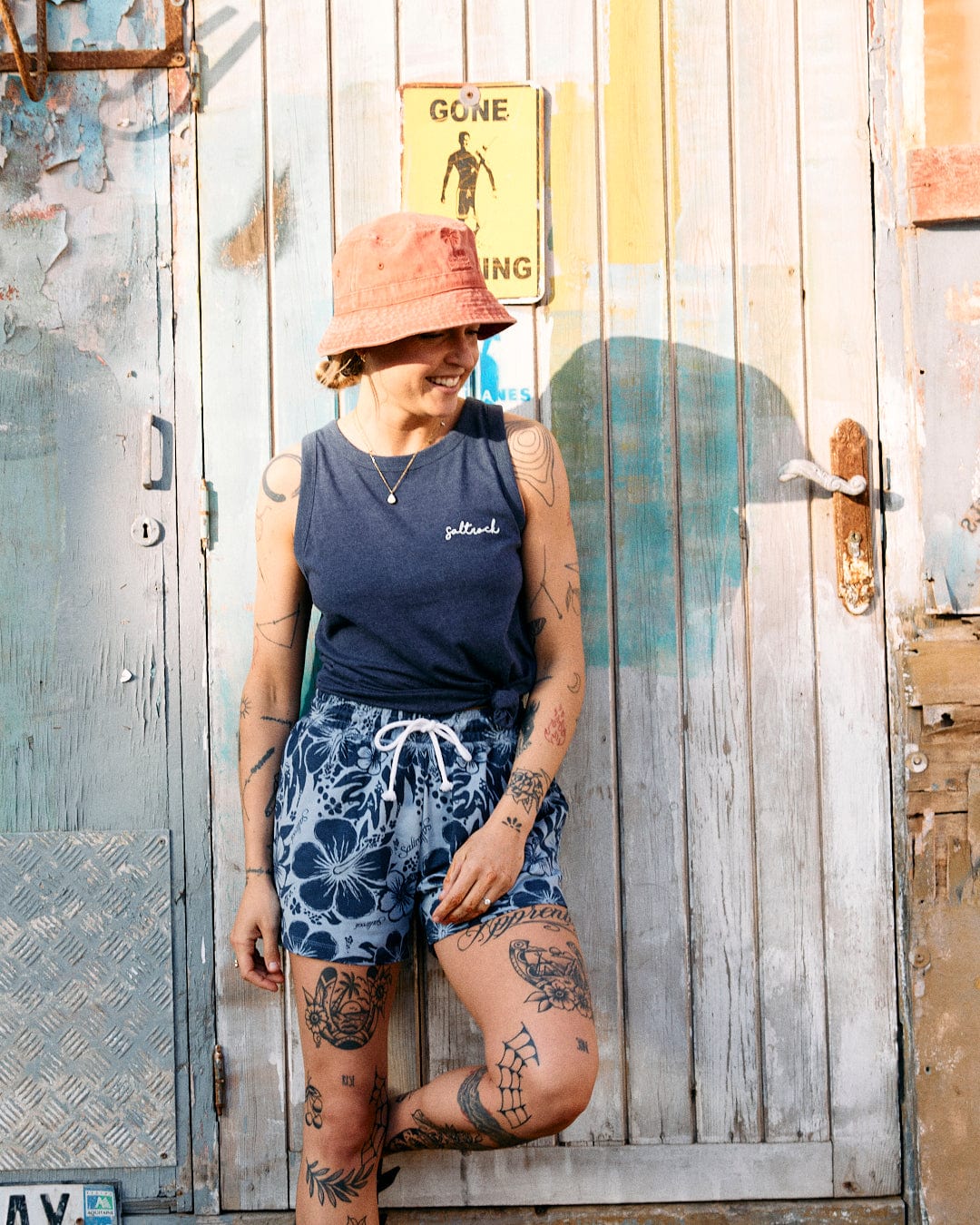 A person with tattoos smiles, standing in front of a rustic door, wearing a pink bucket hat, a Saltrock Velator Womens Vest in Blue with a crew neckline, and floral shorts.