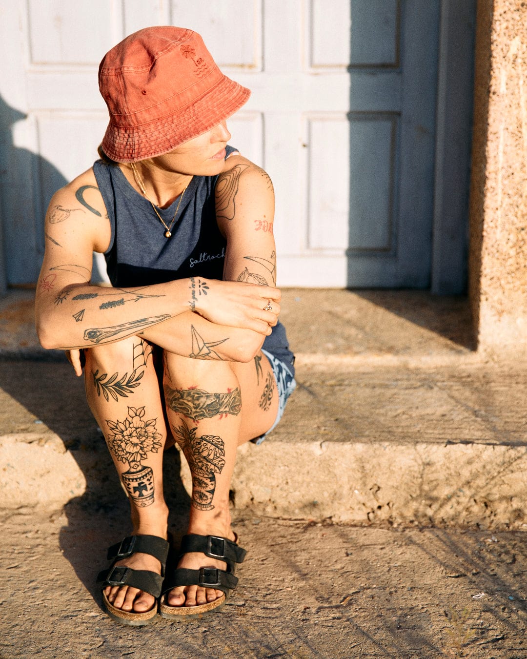 A person with tattoos sits by a wooden door, wearing a red bucket hat and Saltrock machine washable sandals, bathed in sunlight.