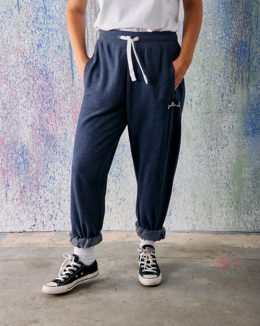 A person wearing Saltrock Velator Women's Joggers in Blue Marl, a white t-shirt, white socks, and black sneakers stands against a textured multicolored wall with hands in pockets.