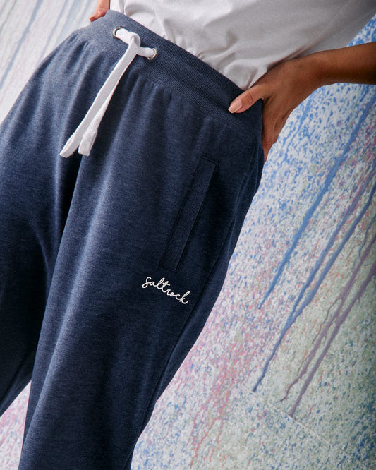A person wears Saltrock Velator Women's Joggers in Blue Marl with an elasticated waist, white drawstring, and pocket embroidery, paired with a white t-shirt, standing against a pastel-textured wall.