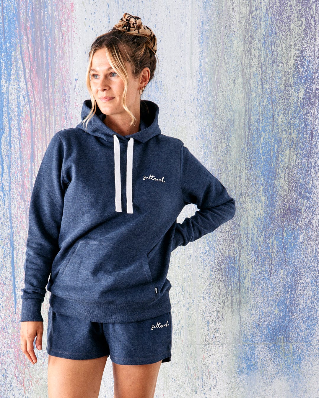 A woman wearing the Saltrock Velator Women's Pop Hoodie in Blue Marl stands in front of a pastel-colored, textured wall.