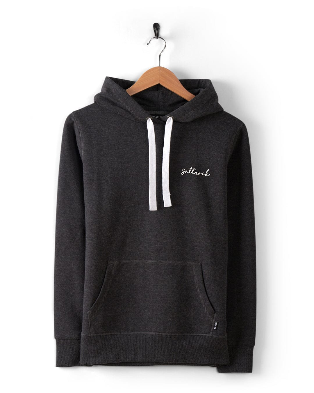 The Saltrock Velator Women's Pop Hoodie in dark grey features white drawstrings, a front pocket, and "saltrock" embroidered on the chest. Made from soft cotton and recycled polyester for comfort and sustainability. Shown on a wooden hanger against a white background.