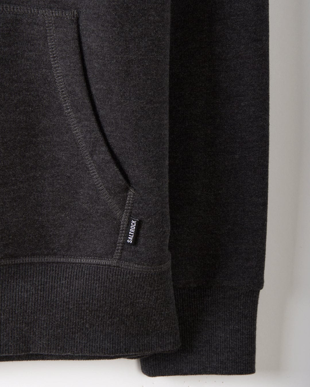 A close-up of the pocket and hem on the Saltrock Velator - Womens Pop Hoodie in dark grey, featuring a small black label near the seam and highlighting its quality craftsmanship and soft, comfortable fabric.
