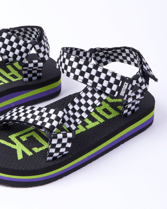 A pair of Saltrock black Warp Time kids sandals with green "pilot" text, checkered Velcro straps, and a purple stripe on a white background.