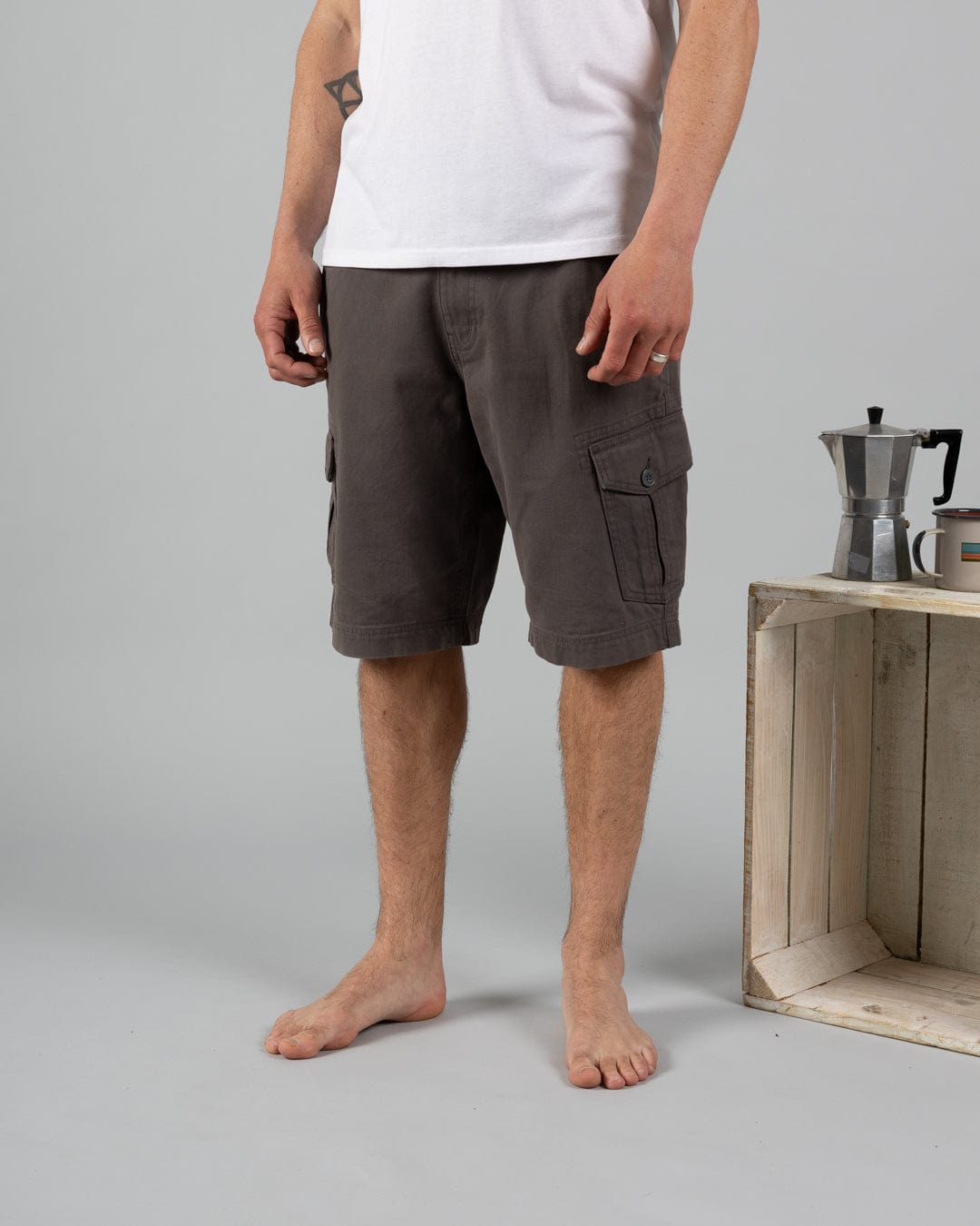 A barefoot person in a white 100% cotton t-shirt and Saltrock’s Penwith II Men's Cargo Shorts in Dark Grey stands by a wooden crate with a moka pot and cup on top.