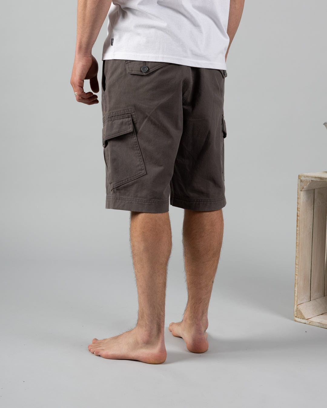 A person stands barefoot on a gray floor, facing away while wearing a white 100% cotton t-shirt and Saltrock Penwith II Men's Cargo Shorts in Dark Grey with patch pockets. A wooden crate on the right adds to the authentic vibe.