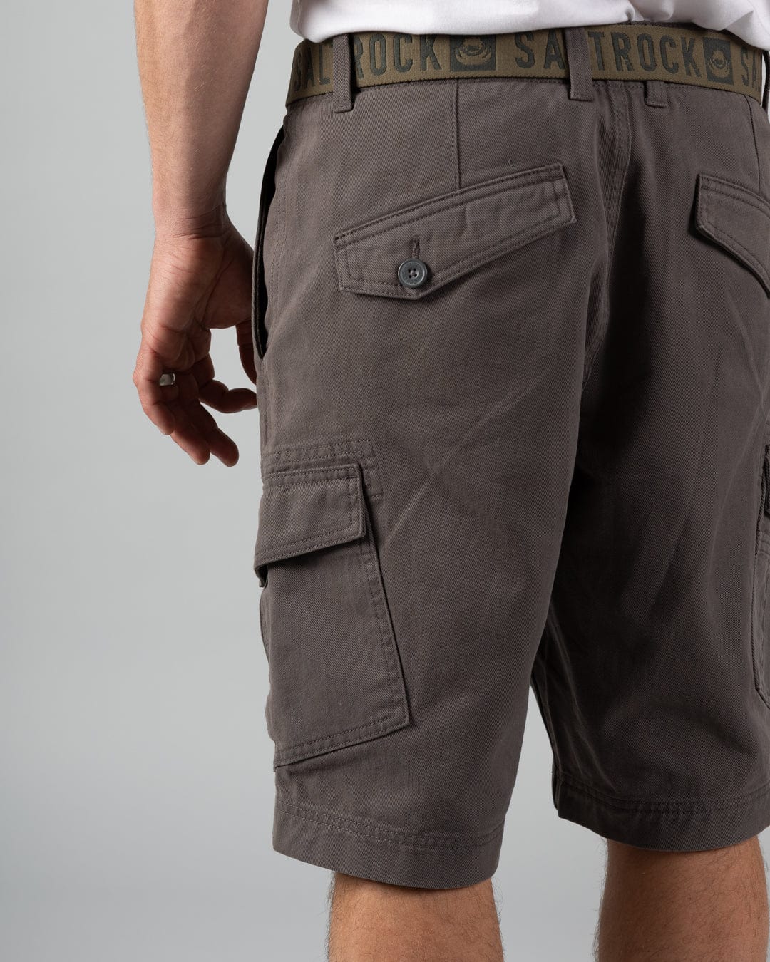 A person models Saltrock's Penwith II Men's Cargo Shorts in dark grey, featuring 100% cotton fabric and buttoned cargo patch pockets, styled with a black belt and photographed waist down against a plain backdrop.
