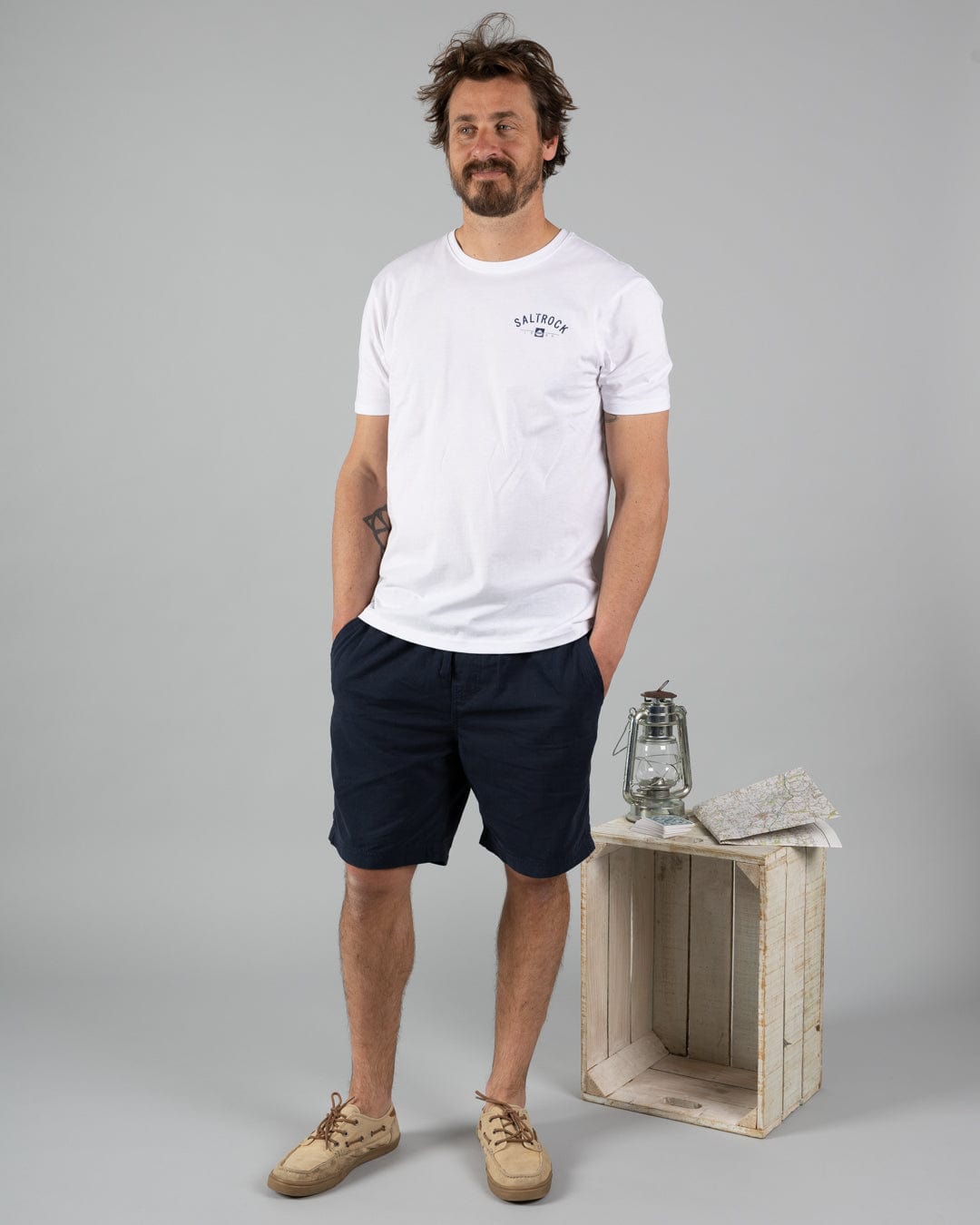 A man stands with hands in pockets, wearing a white T-shirt, Saltrock Meddon Men's Chino Shorts in blue, and beige shoes. Next to him is a wooden crate topped with a lantern and a folded map.