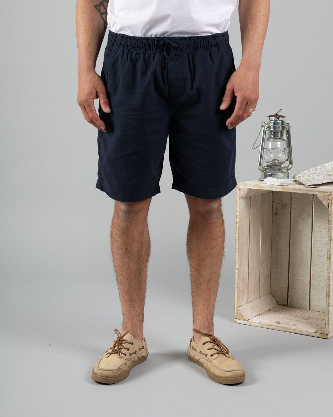 A person wearing a white t-shirt, Saltrock Meddon Mens Chino Shorts in blue, and beige shoes stands next to a wooden crate topped with a lantern and a closed book.