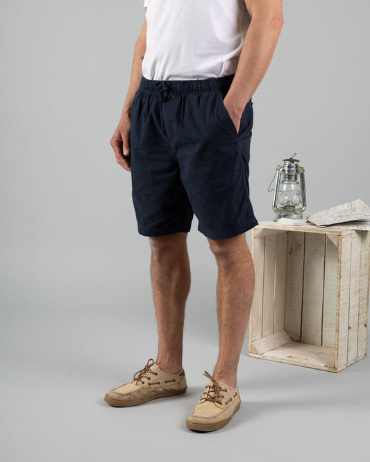 A man in a white t-shirt, Saltrock Meddon Men's Chino Shorts in blue, and beige sneakers stands beside a white wooden crate topped with a lantern and folded map.