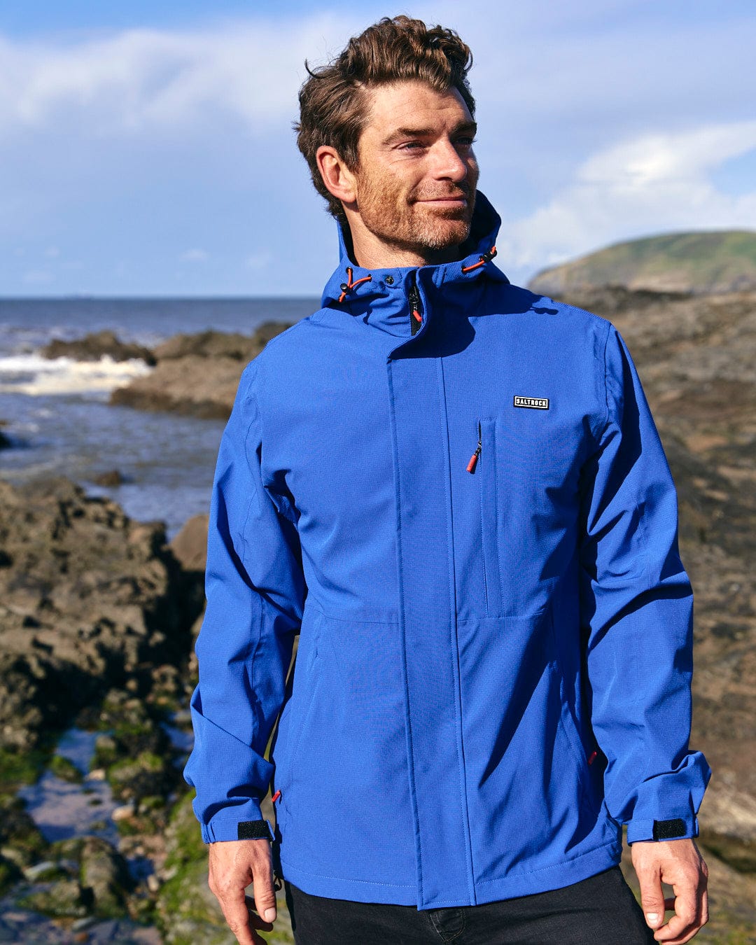 A man in a Saltrock Whistler Mens Hooded Jacket in Blue standing on rocks near the ocean.