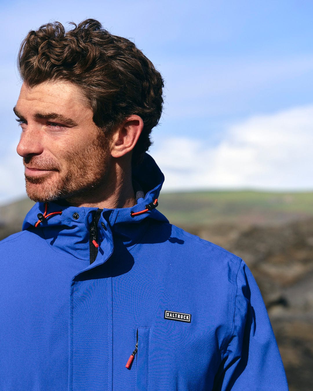 A man in a Saltrock Whistler - Mens Hooded Jacket - Blue is standing in front of rocks.