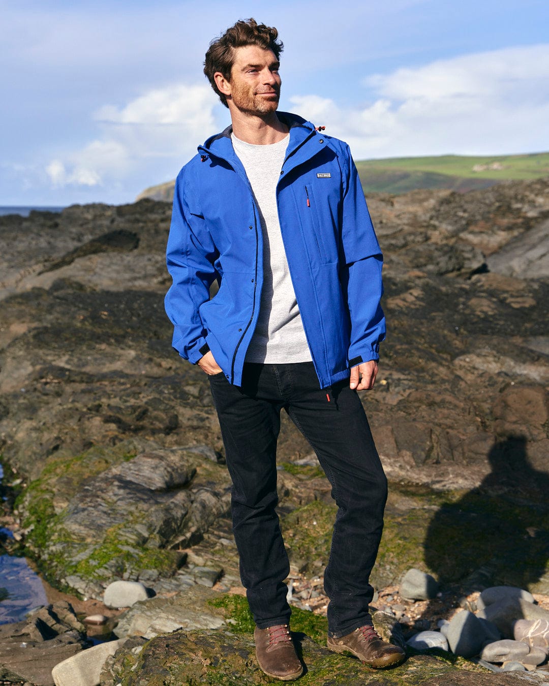 A man in a Saltrock Whistler - Mens Hooded Jacket - Blue, waterproof and standing on a rocky beach.
