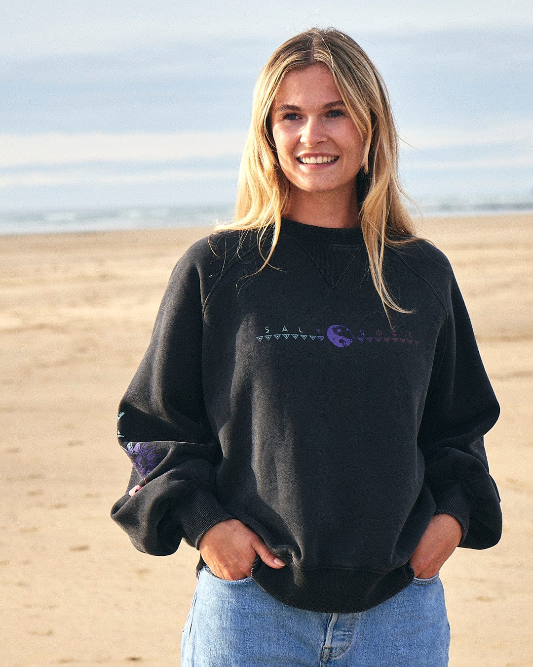A woman wearing a Winter Solstice Gradient - Womens Sweat - Washed Black sweatshirt by Saltrock on the beach.