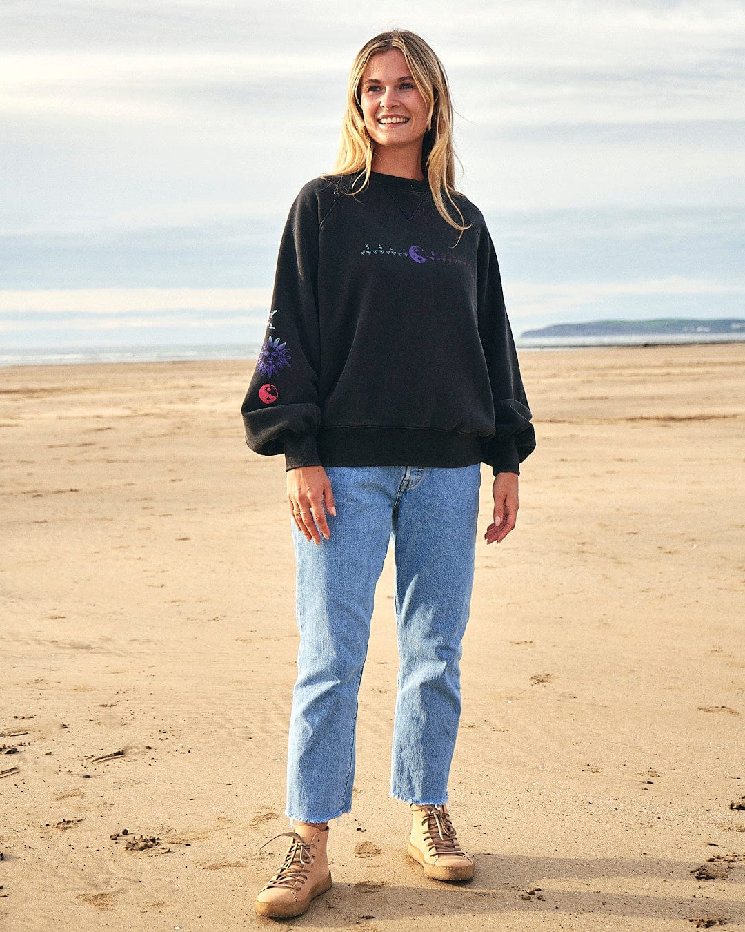 A woman wearing a Saltrock Winter Solstice Gradient - Womens Sweat - Washed Black sweatshirt standing on the beach.