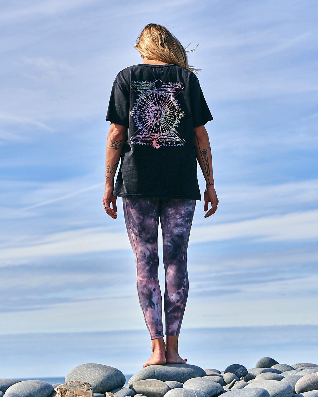 A woman wearing a Saltrock Winter Solstice Gradient - Womens Short Sleeve T-Shirt - Washed Black and leggings standing on rocks.