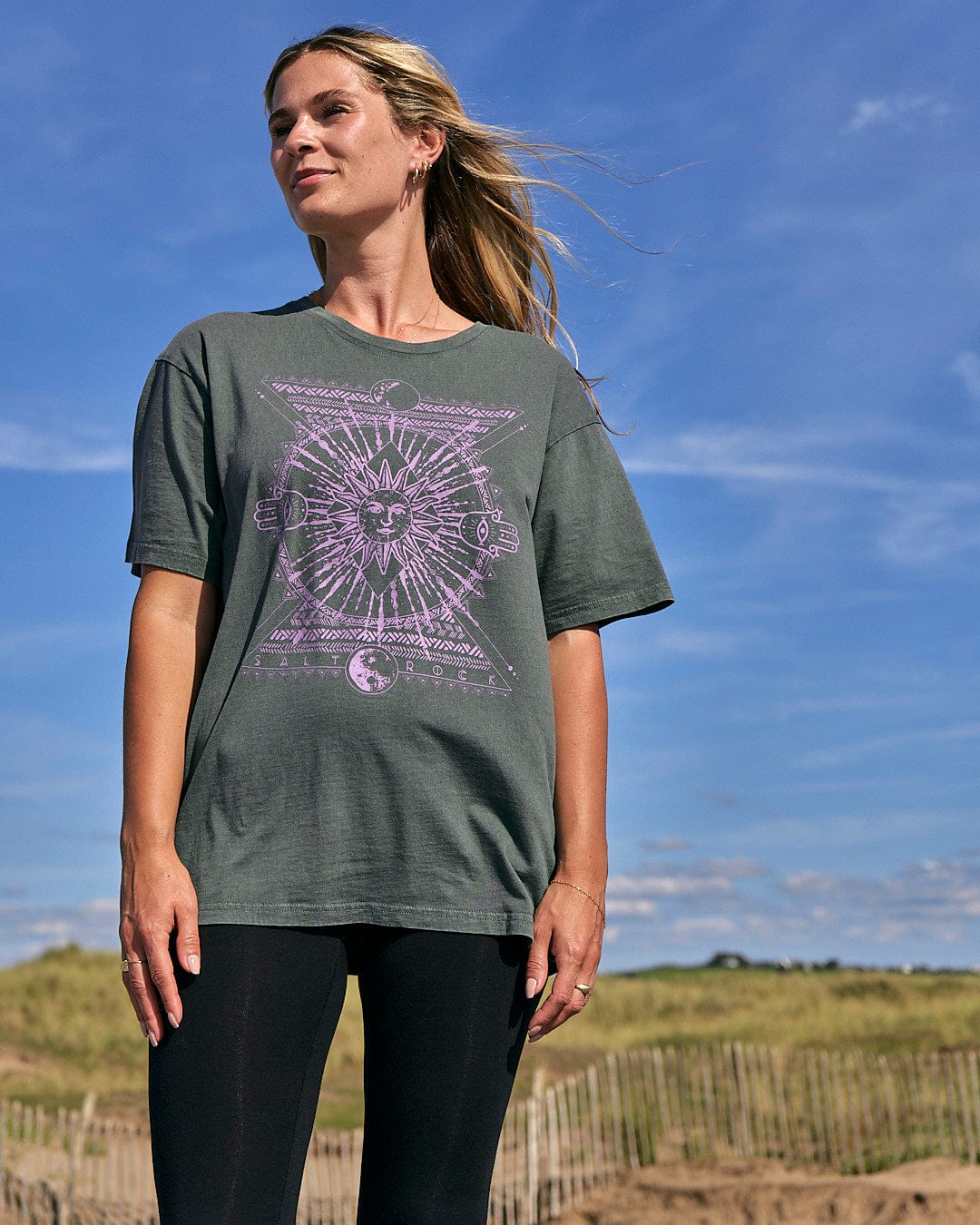 A woman wearing a purple graphic Saltrock Winter Solstice - Womens Short Sleeve T-Shirt - Dark Green standing on the beach.