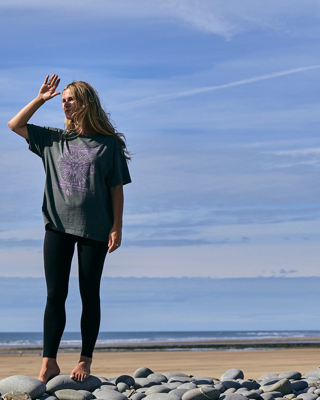 A woman wearing a 100% cotton Saltrock Winter Solstice - Womens Short Sleeve T-Shirt - Dark Green, standing on rocks on the beach.