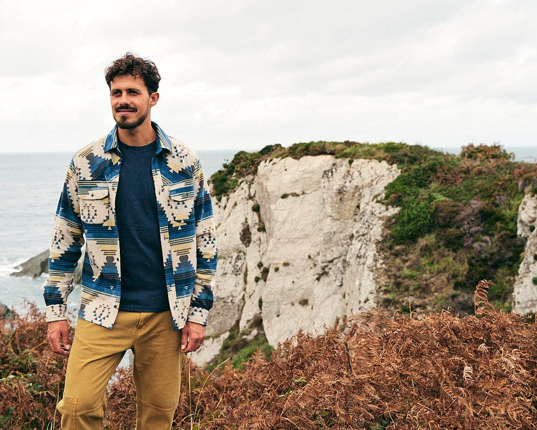 A man wearing a Saltrock Wolfe - Mens Aztec Fleece Shirt - Blue stands on top of a cliff, gazing at the vast ocean with a sense of comfort and warmth.