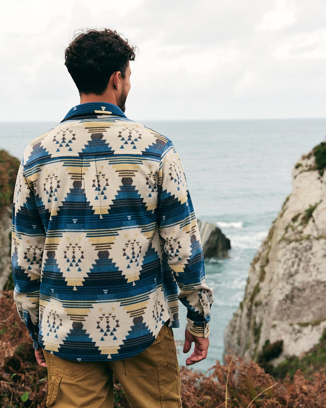 A man is standing on a cliff, feeling the warmth of his Saltrock - Wolfe - Mens Aztec Fleece Shirt - Blue as he gazes at the vast ocean.