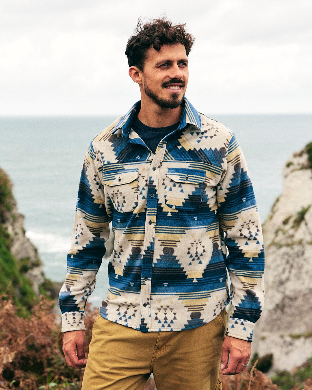 A man wearing a Saltrock Wolfe - Mens Aztec Fleece Shirt - Blue standing by the ocean.