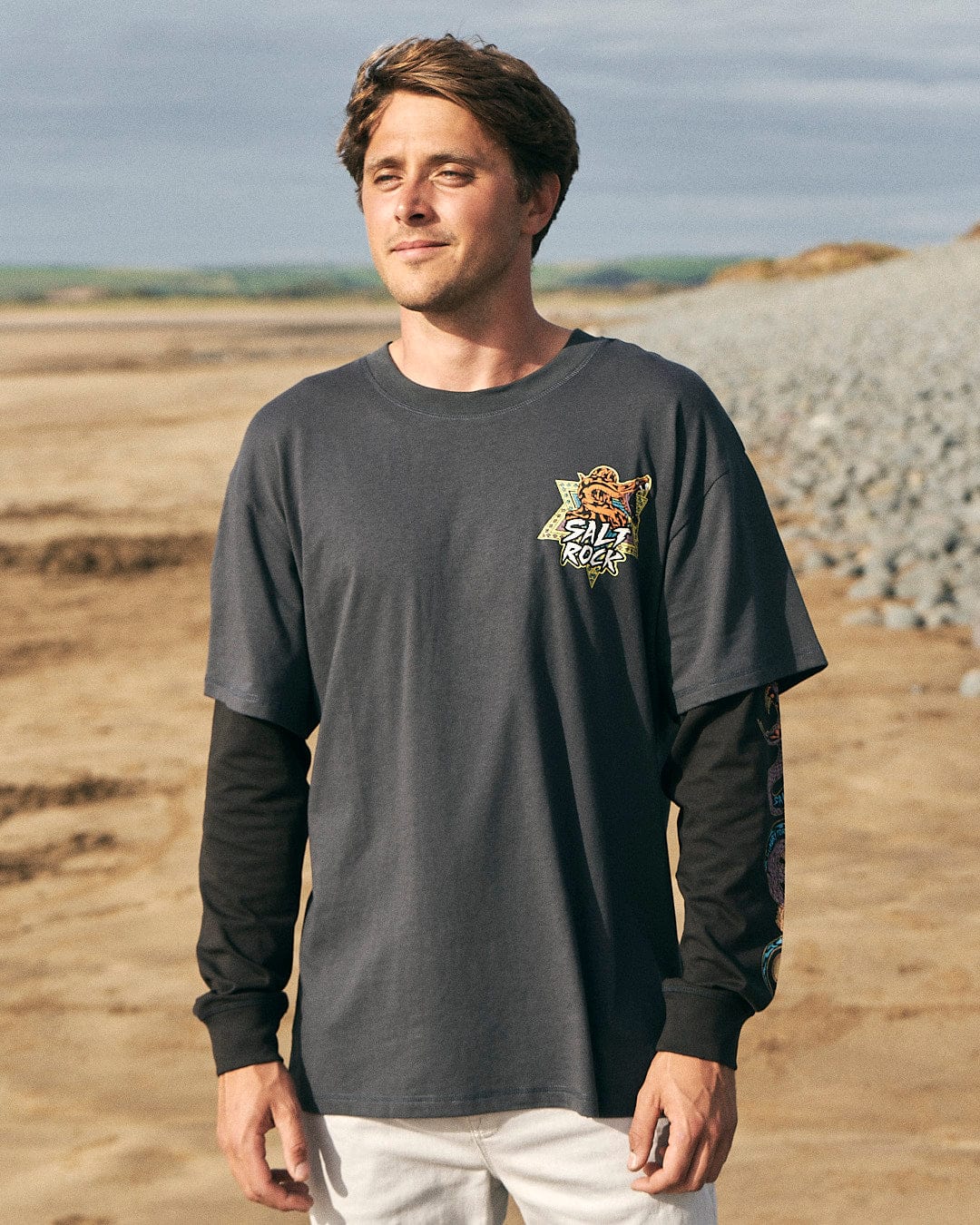 A man wearing a Saltrock Wolfscale - Mens Double Layer Long Sleeve T-Shirt - Black standing on a beach.