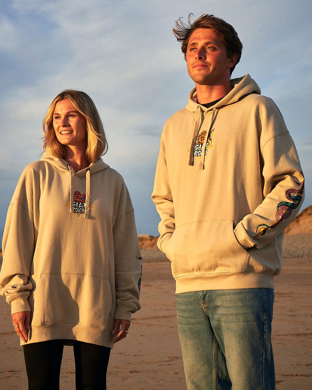 A man and woman standing on a beach wearing Saltrock hoodies with Wolfscale - Pop Hoodie - Beige print branding.