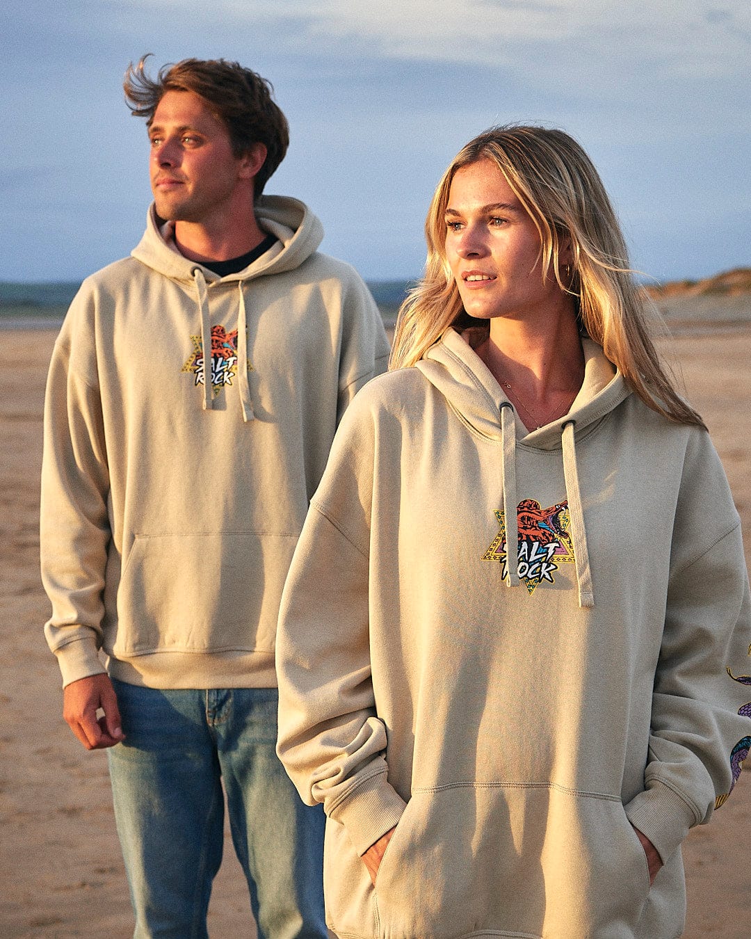 A man and woman standing on a beach wearing Saltrock's Wolfscale - Pop Hoodie - Beige.