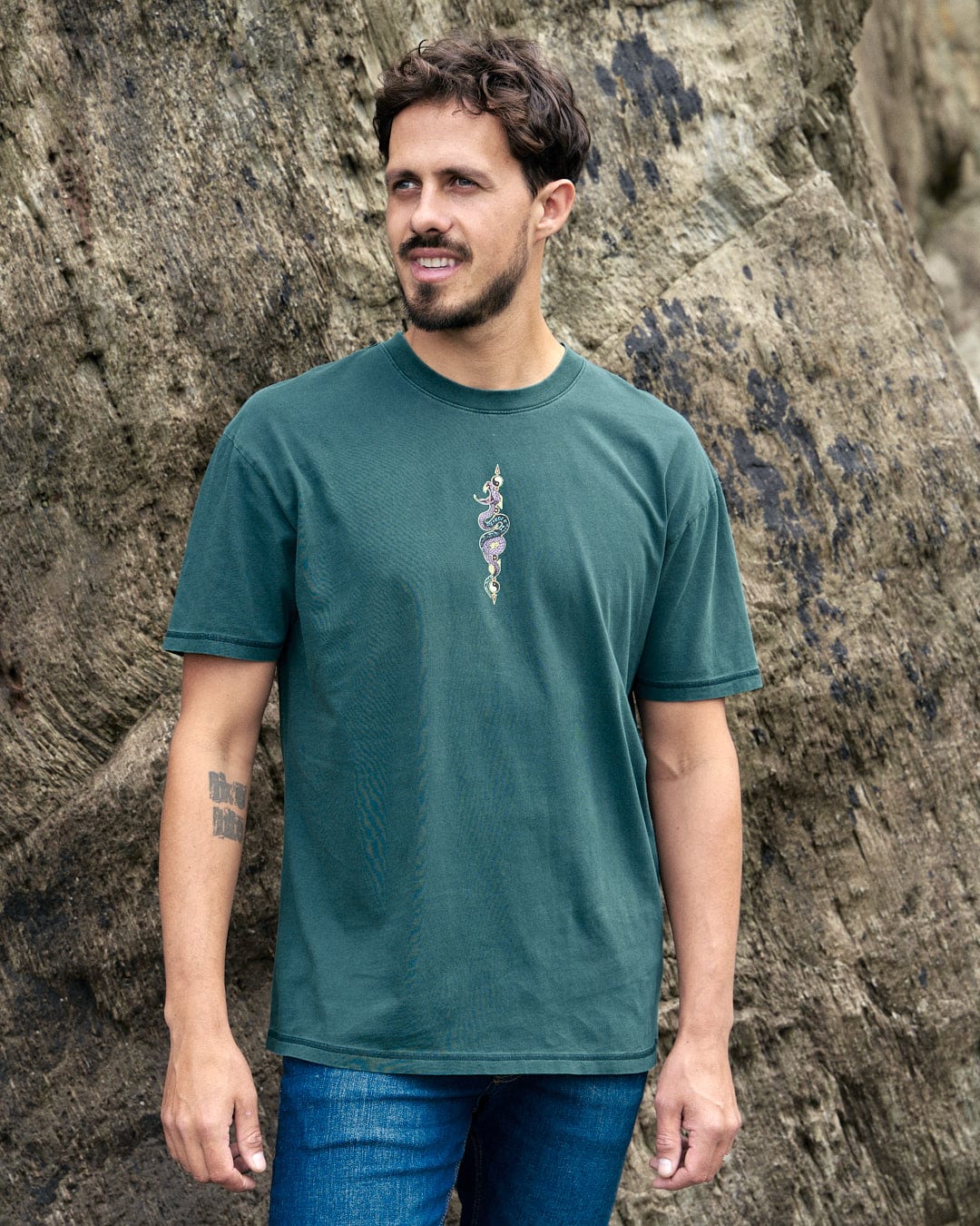 A man wearing a Saltrock Wolfscale Mens Short Sleeve T-Shirt in Dark Green, standing in front of rocks.