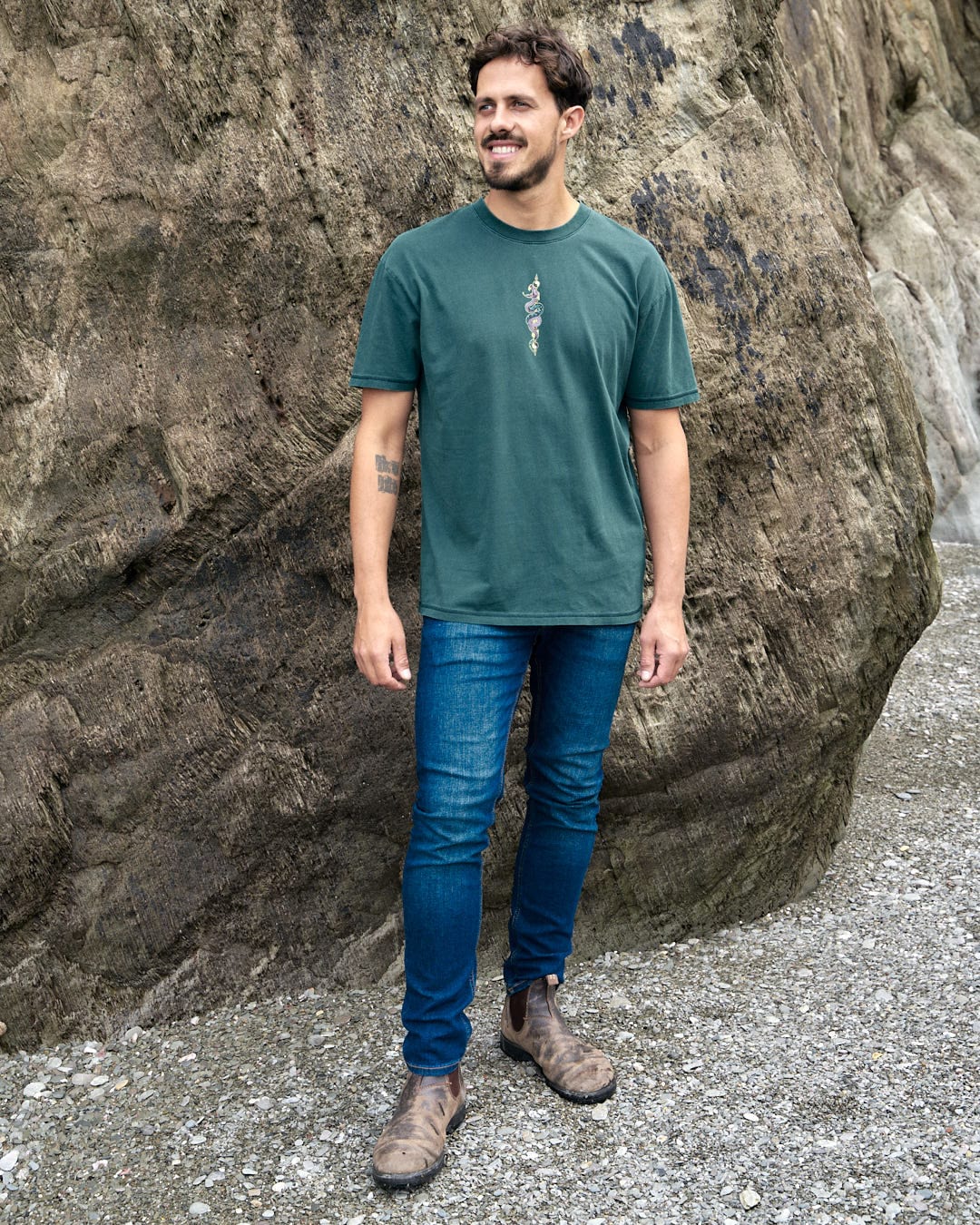 A man in a Saltrock Wolfscale - Mens Short Sleeve T-Shirt - Dark Green standing in front of rocks, wearing a graphic snake print.