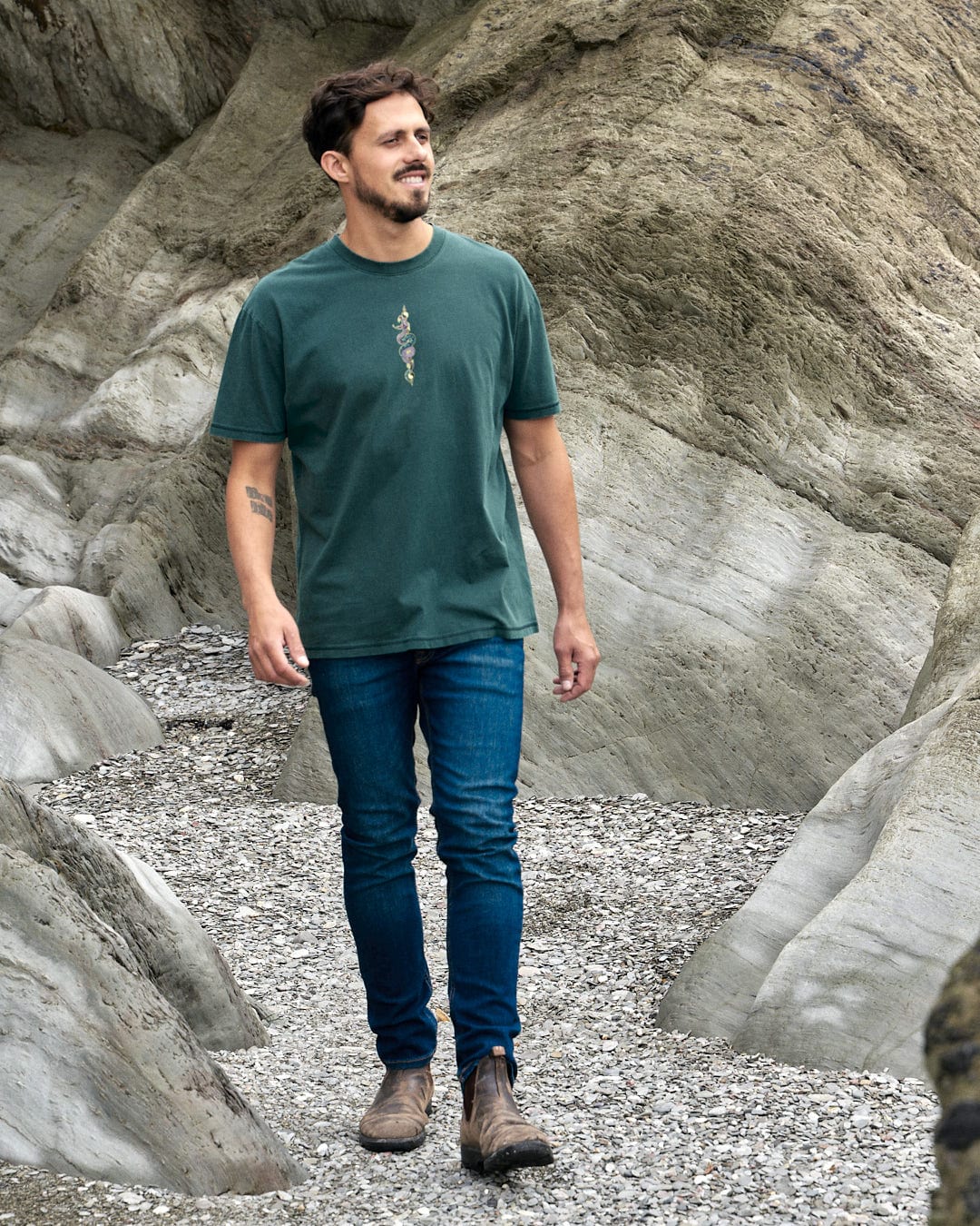 A man wearing a Saltrock Wolfscale - Mens Short Sleeve T-Shirt - Dark Green walking on rocks with a snake graphic.