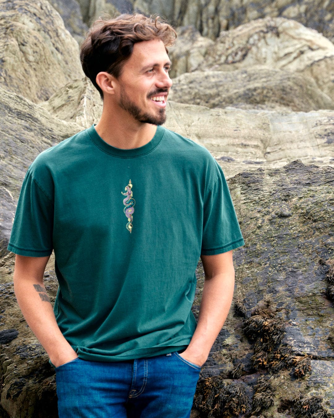 A man wearing a Saltrock Wolfscale - Mens Short Sleeve T-Shirt - Dark Green with a snake graphic in front of rocks.