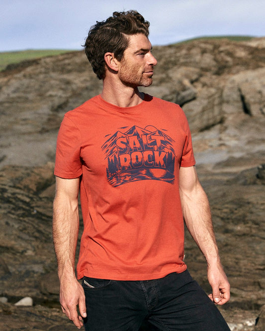 A stylish man wearing a Wood Carve Logo - Mens Short Sleeve T-Shirt - Red by Saltrock is standing on rocks.