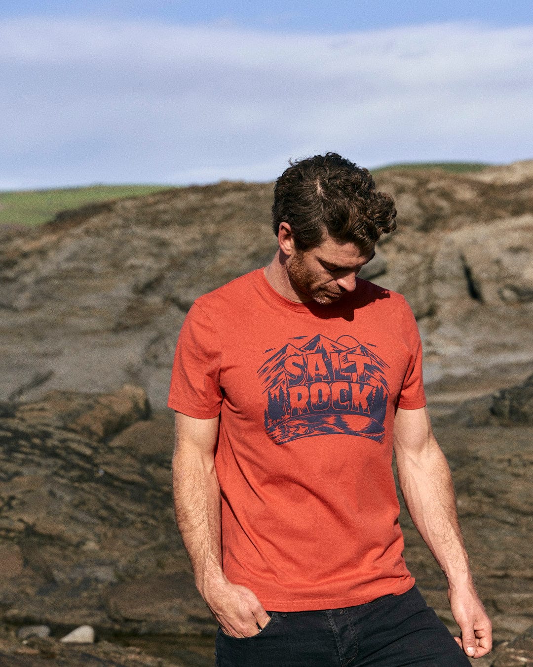 A stylish man wearing a Saltrock Wood Carve Logo - Mens Short Sleeve T-Shirt - Red standing on rocks in a mountain scene.