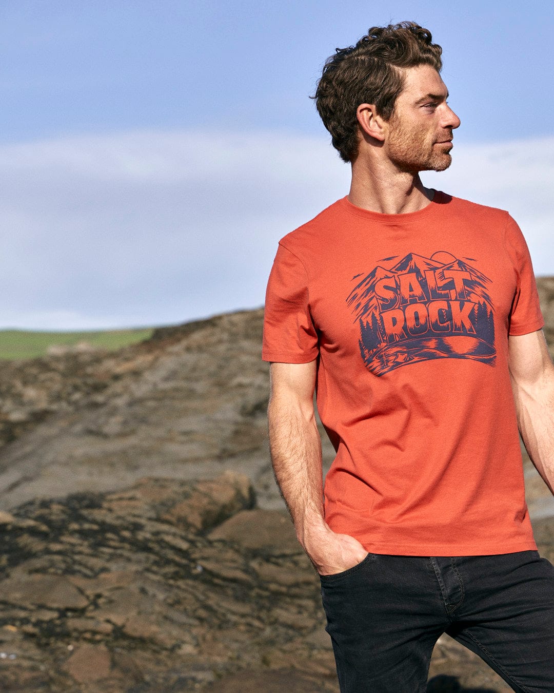 A stylish man wearing a Saltrock Wood Carve Logo - Mens Short Sleeve T-Shirt - Red standing on a rock amidst a picturesque mountain scene.